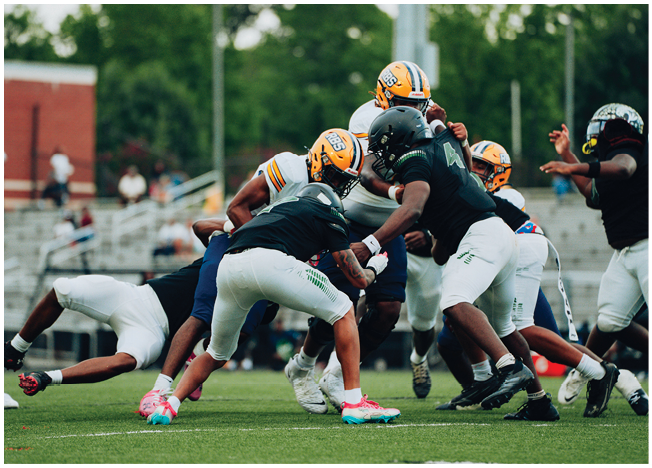 Several people wearing football helmets and uniforms are engaged in a play on a grassy field with a blurred background of trees and bleachers. Description generated by AI