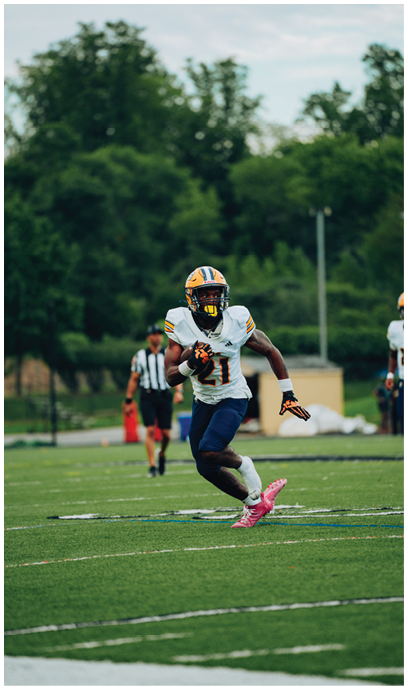 A person wearing a football uniform with a helmet and pink cleats is running on a grassy field while holding a football. Description generated by AI