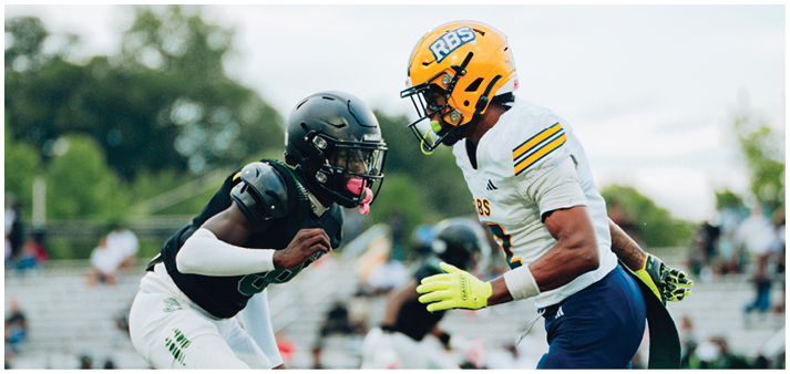 Two people wearing football helmets and uniforms are facing each other on a field, appearing to be in the middle of a play or practice. Description generated by AI