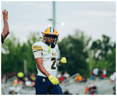 A person wearing a yellow helmet, white football jersey with the number 2, and blue pants stands on a football field with blurred spectators in the background. Description generated by AI
