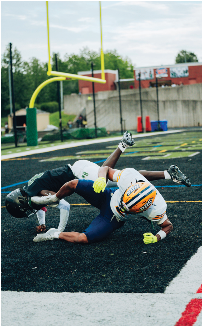 Two people wearing football gear are tangled on the ground during a game on a black turf field near the end zone. Description generated by AI
