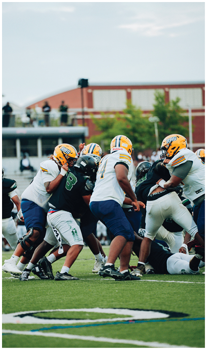 Several people wearing football helmets and uniforms are engaged in a play on a grassy field with a building and spectators in the background. Description generated by AI