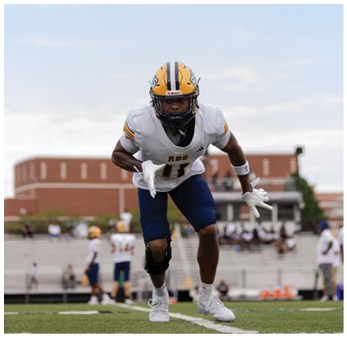 A person wearing a football uniform and helmet is running forward on a field with a blurred background of other players and bleachers. Description generated by AI