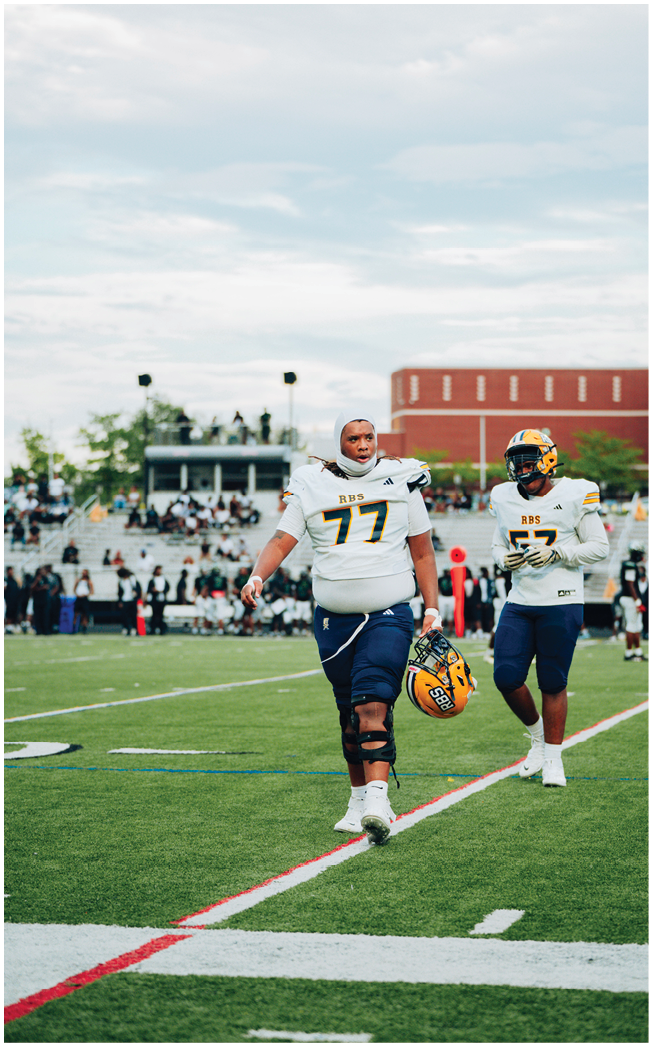 Two people in football uniforms are walking on a field with one holding a helmet and wearing a knee brace. Description generated by AI