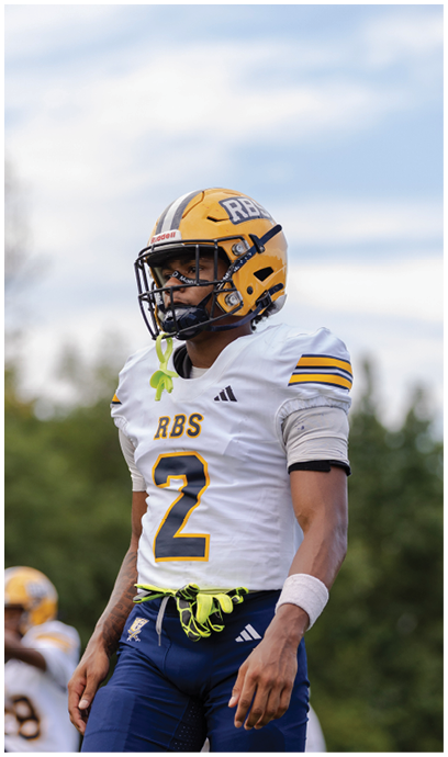 A person wearing a yellow football helmet and white jersey with the number 2 stands on a field with trees and a cloudy sky in the background. Description generated by AI