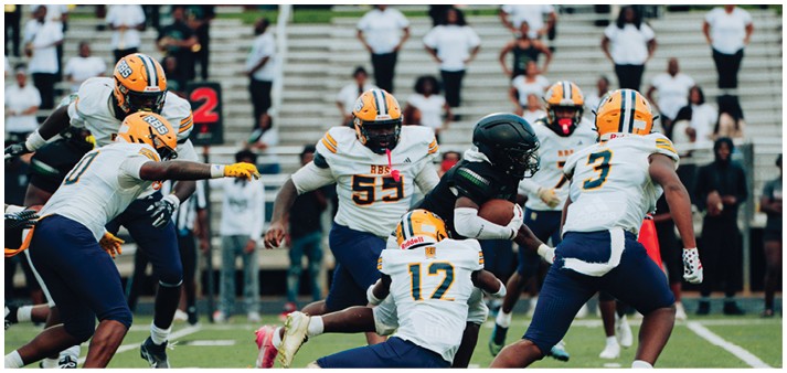 A group of people in football uniforms are actively engaged in a play on a field with one person holding the ball while others attempt to block or tackle. Description generated by AI