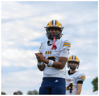 A person wearing a yellow and white football uniform with a helmet stands on a field with another similarly dressed person in the background under a cloudy sky. Description generated by AI