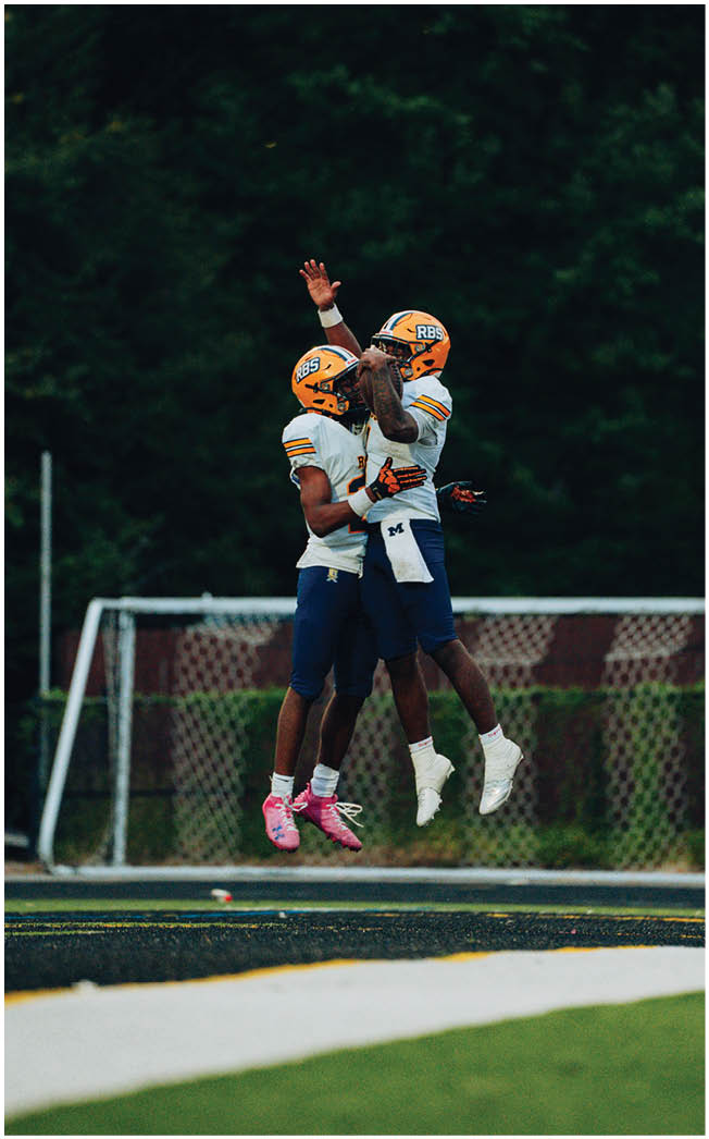 Two people in football uniforms jump and celebrate together on a field near a goalpost with trees in the background. Description generated by AI