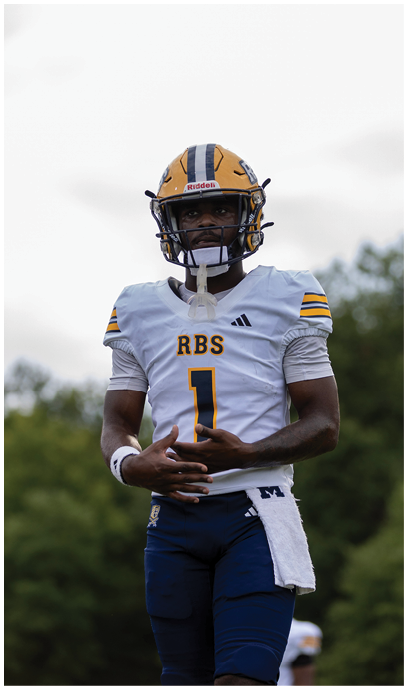 A person wearing a yellow and blue football helmet and a white football jersey with the number one and the letters RBS stands outdoors with trees in the background. Description generated by AI