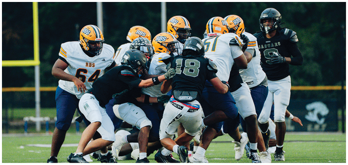 A group of people wearing football helmets and uniforms are engaged in a play on a grassy field with goalposts and trees in the background. Description generated by AI