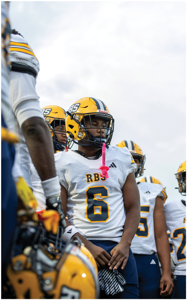 A group of people wearing yellow helmets and white football jerseys with navy pants are standing together, with one person in the center holding gloves and a pink mouthguard hanging from their helmet. Description generated by AI