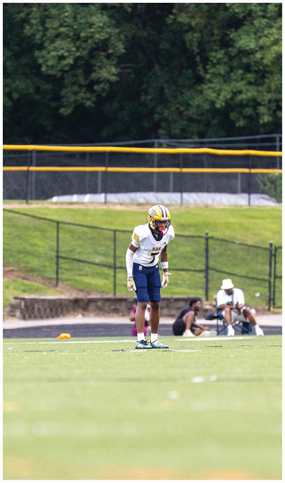 A person in a football uniform stands on a grassy field with trees and a fence in the background while two people sit on the ground near the fence. Description generated by AI