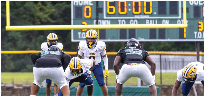 A group of people in football uniforms are positioned on a field ready for a play with a scoreboard showing the time in the background. Description generated by AI