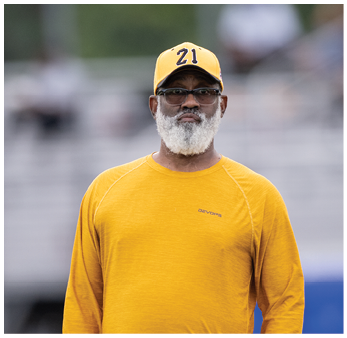 A person wearing a yellow long-sleeve shirt and brown pants stands on a grassy field with hands in pockets. Description generated by AI