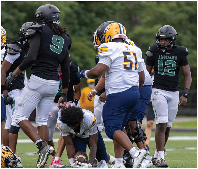People wearing football uniforms are gathered on a field with one person kneeling and holding a football while others stand nearby. Description generated by AI