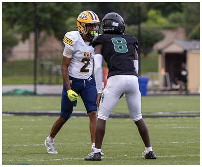 Two people wearing football uniforms are facing each other on a grassy field with trees and a building in the background. Description generated by AI