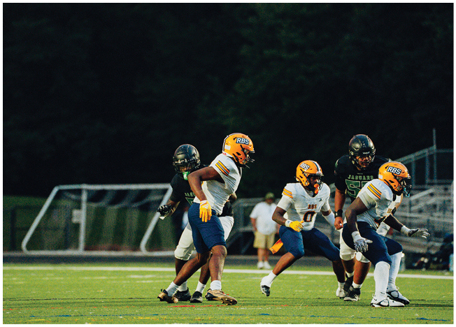 Four people wearing football uniforms and helmets are engaged in a play on a grassy field with a goalpost and bleachers in the background. Description generated by AI