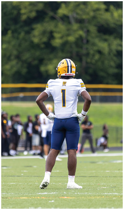 A person wearing a yellow and blue football helmet, white jersey with the number one, and blue pants stands on a football field with hands on hips facing away from the camera. Description generated by AI