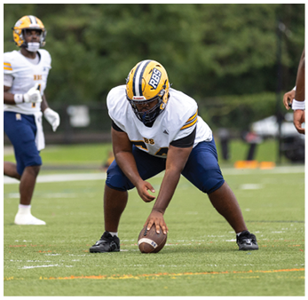A person in a football uniform is crouched on a field holding a football, preparing to snap it, while another person in similar attire stands in the background. Description generated by AI