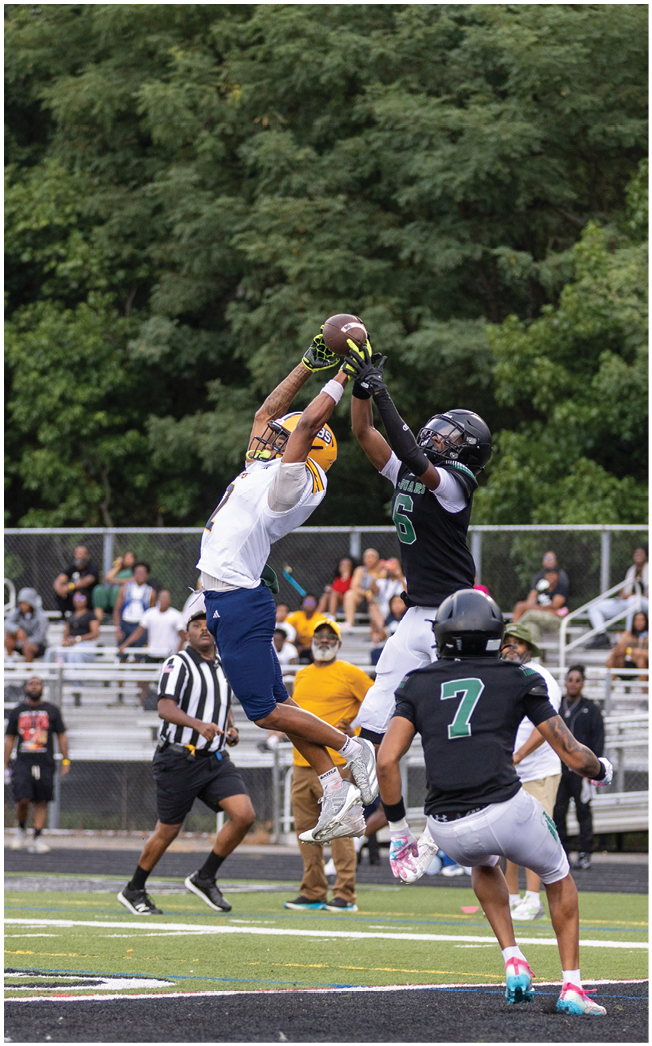 Two football players in mid-air compete to catch the ball while another player and a referee watch on a field with spectators in the background. Description generated by AI
