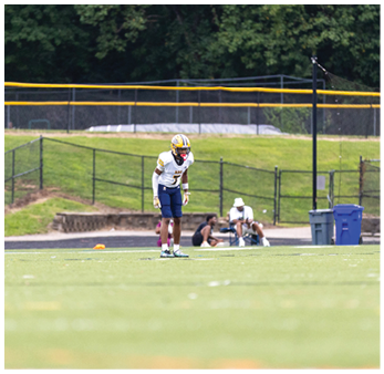 A person in a football uniform stands on a field with a helmet and cleats while two people sit in the background near a fence and trash bins. Description generated by AI
