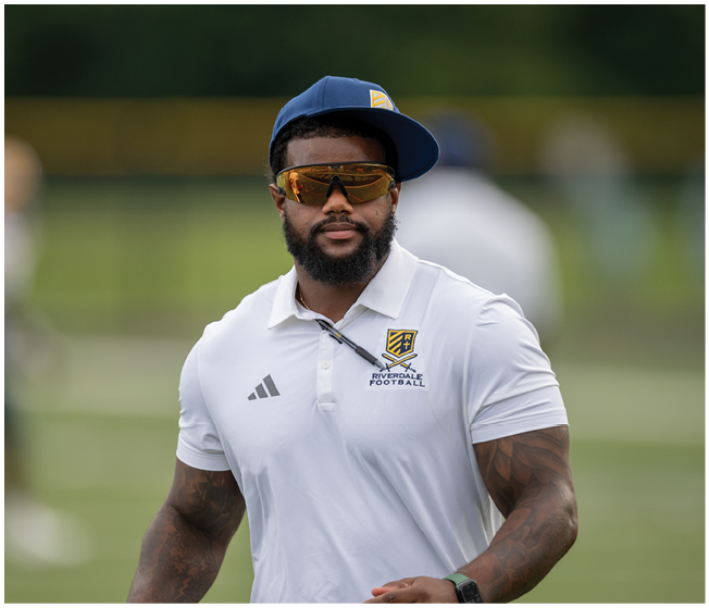A person wearing a white Adidas polo shirt with a sports team logo is standing outdoors on a grassy field. Description generated by AI