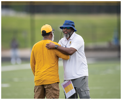 Two people stand on a sports field wearing casual clothing, with one holding a clipboard. Description generated by AI