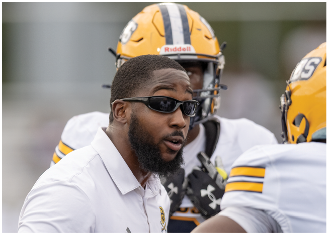 A person wearing sunglasses and a white polo shirt with a logo is speaking to another person in a white sports jersey with yellow and black stripes on the sleeves. Description generated by AI