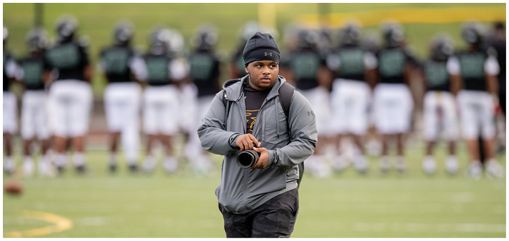 A person wearing a beanie and gray jacket is holding a camera and walking on a sports field with a blurred group of people in sports uniforms standing in the background. Description generated by AI