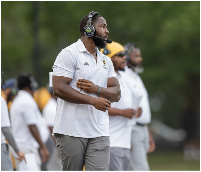 A person wearing a white polo shirt and headset stands on a field with two other people in similar attire blurred in the background. Description generated by AI