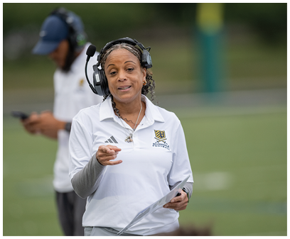 A person wearing a headset and a white sports polo shirt is holding a clipboard and speaking on a sports field with another person in the background looking at a phone. Description generated by AI