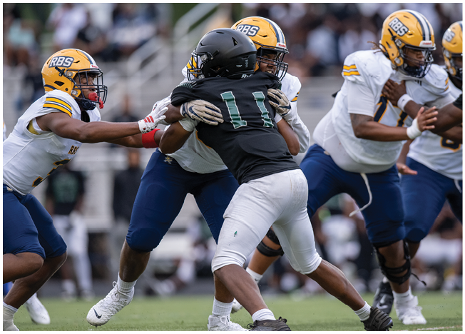 People wearing football uniforms are engaged in a close physical play on a grassy field during a game. Description generated by AI