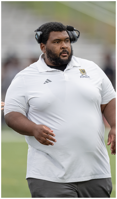 Person wearing a white polo shirt and headset standing on a sports field. Description generated by AI