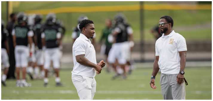 Two adults wearing white shirts and light-colored pants are standing and talking on a sports field with a group of people in sports uniforms blurred in the background. Description generated by AI
