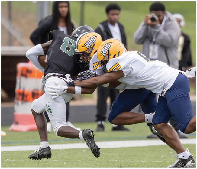 Two people in yellow helmets and white jerseys tackle a person in a black jersey and white pants during a football game on a grassy field. Description generated by AI