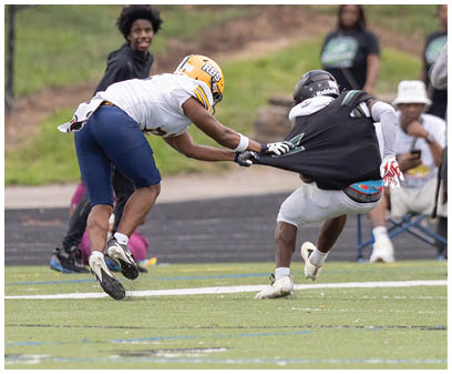 Two people in football gear are engaged in a play on a field with one person pulling the other's jersey while several people watch and one takes a photo in the background. Description generated by AI