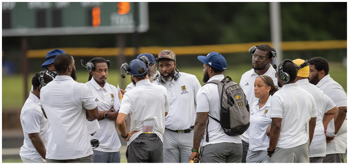 A group of people wearing white shirts and headsets are gathered in a circle on a sports field, engaged in discussion. Description generated by AI