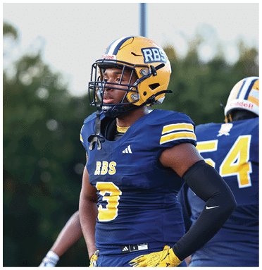 A person wearing a yellow football helmet and blue uniform with the number 3 stands on a field with another player in the background. Description generated by AI