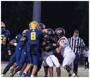 Players in blue and white football uniforms are engaged in a play on a field with a dark background. Description generated by AI