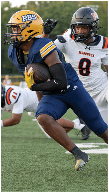 A person in a yellow helmet and blue football uniform is running with a football while another person in a white football uniform with the number 9 is attempting to catch them on a grassy field. Description generated by AI