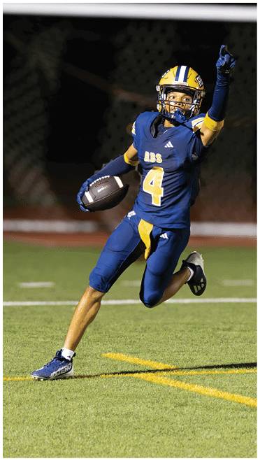 A person wearing a blue and yellow football uniform with the number four is holding a football and appears to be preparing to throw it during a game. Description generated by AI