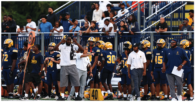 A group of people in sports uniforms and coaches stand on the sidelines of a football field with spectators visible in the background. Description generated by AI