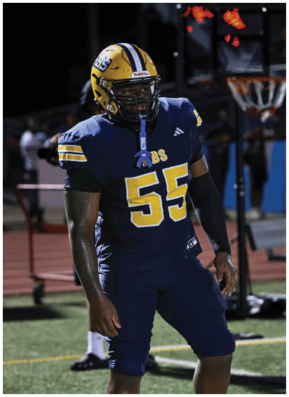 A person wearing a blue and yellow football uniform with the number 55 stands on a field at night near a basketball hoop. Description generated by AI