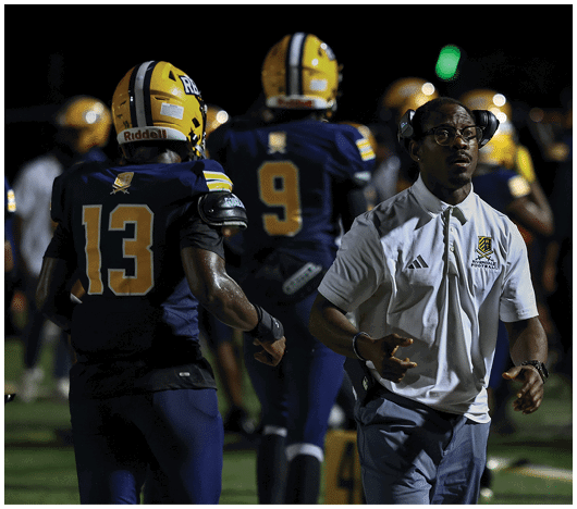 A person wearing a white polo shirt and glasses is standing on a football field near players in blue and yellow uniforms during a nighttime game. Description generated by AI