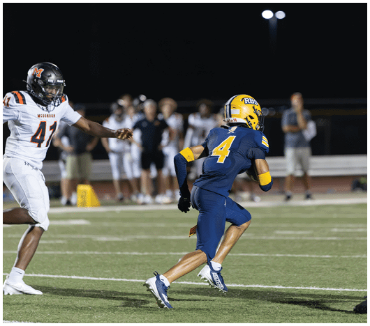 An adult in a blue and yellow football uniform with the number 4 is running on a field while another adult in a white and black uniform with the number 41 is reaching out toward them during a nighttime game. Description generated by AI