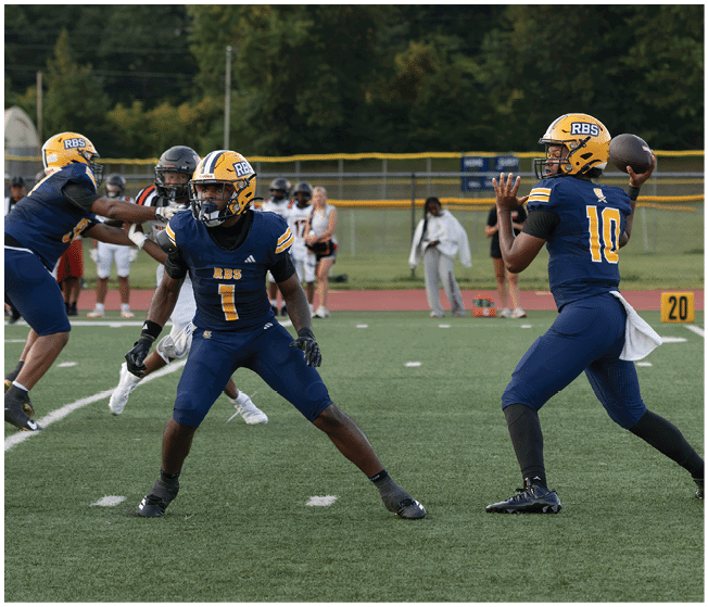 Two people in blue and yellow football uniforms are on a field with one preparing to throw a football and the other positioned in front of them. Description generated by AI