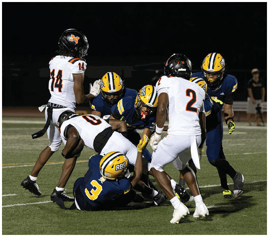 Several people in football uniforms are engaged in a tackle on a grassy field during a nighttime game. Description generated by AI