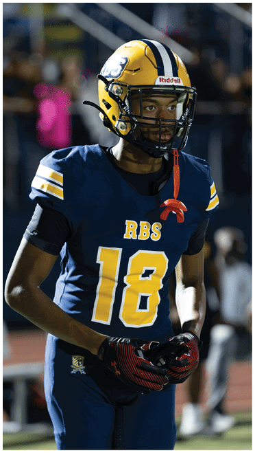 A person wearing a blue and yellow football uniform with the number 18 and a yellow helmet is standing on a field. Description generated by AI