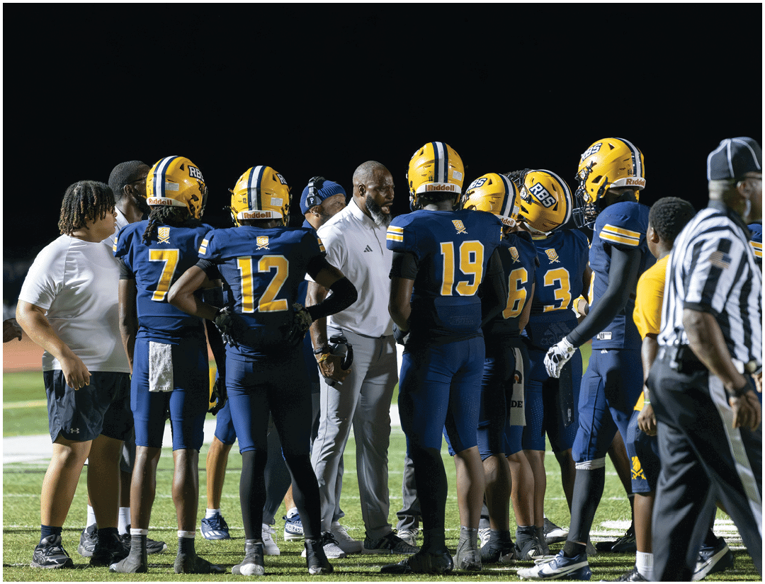 A group of football players in blue and yellow uniforms are gathered around a coach on a field at night while a referee walks nearby. Description generated by AI