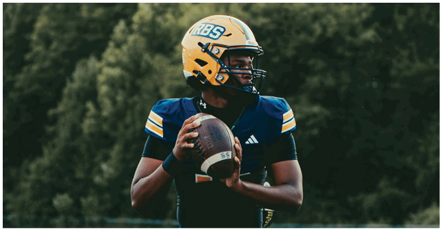 A person wearing a yellow football helmet and dark blue uniform is holding a football and preparing to throw it on a field with trees in the background. Description generated by AI
